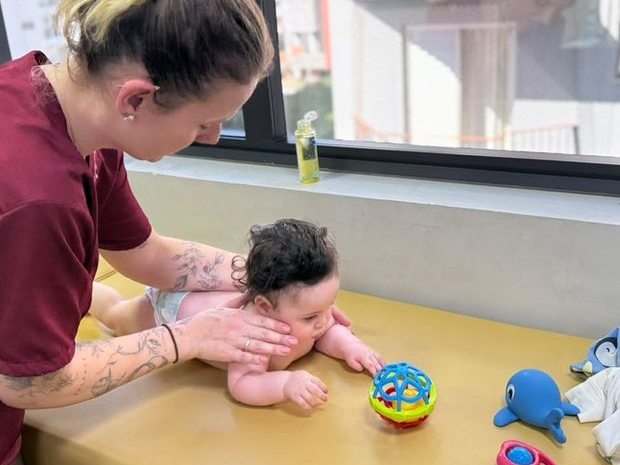 Bebê em decúbito ventral apoiado nos antebraços, alcançando brinquedos sobre a mesa
